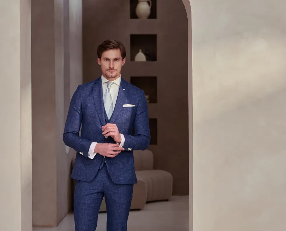 Bruidegom in een strak blauw trouwpak poseert in een modern, minimalistisch interieur met warme aardetinten.