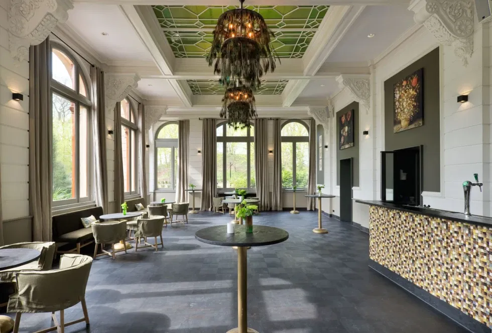 Luxe ontvangstruimte in Oranjerie Hydepark met hoge ramen, stijlvolle bar en groen glas-in-lood plafond – ideale setting voor bruiloftsreceptie of borrel.