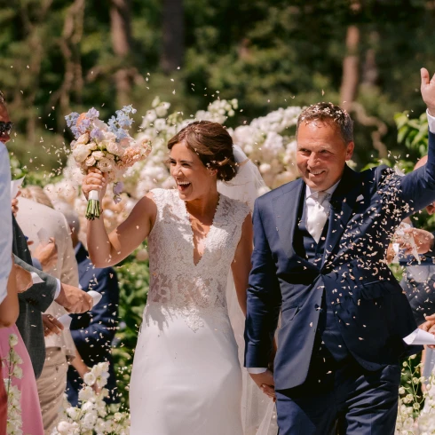 Stralende bruid en bruidegom lopen hand in hand door een bloemenpad terwijl gasten confetti gooien.