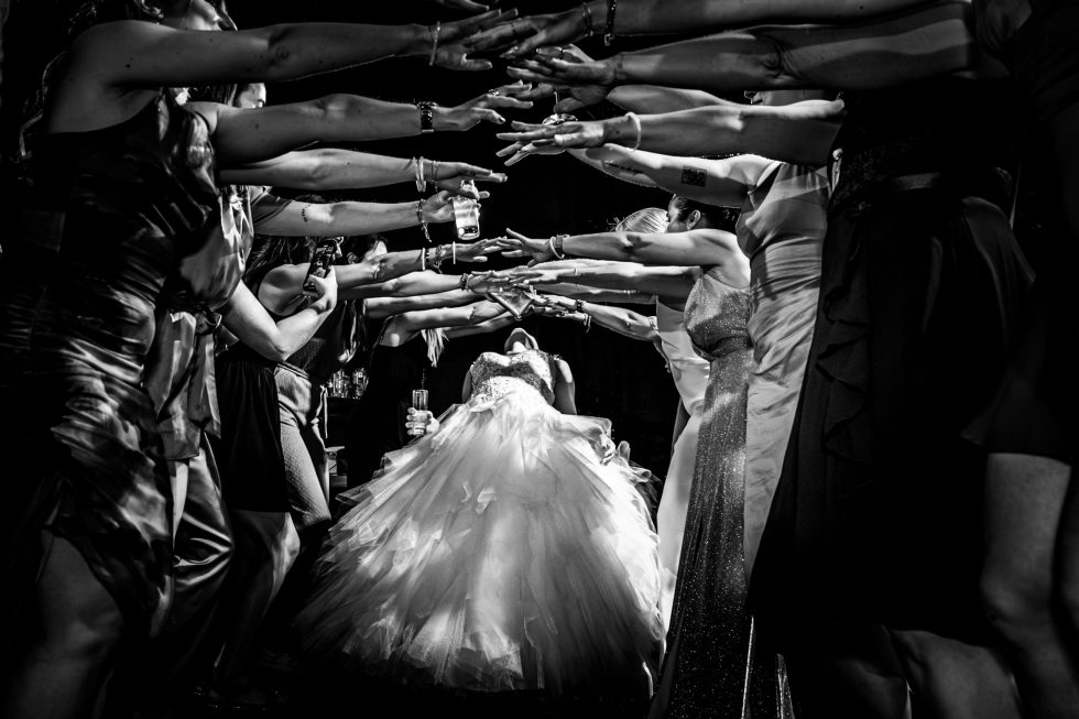 Bruid danst de limbo met een groep vrienden, jurk in beweging, omringd door feestgangers.