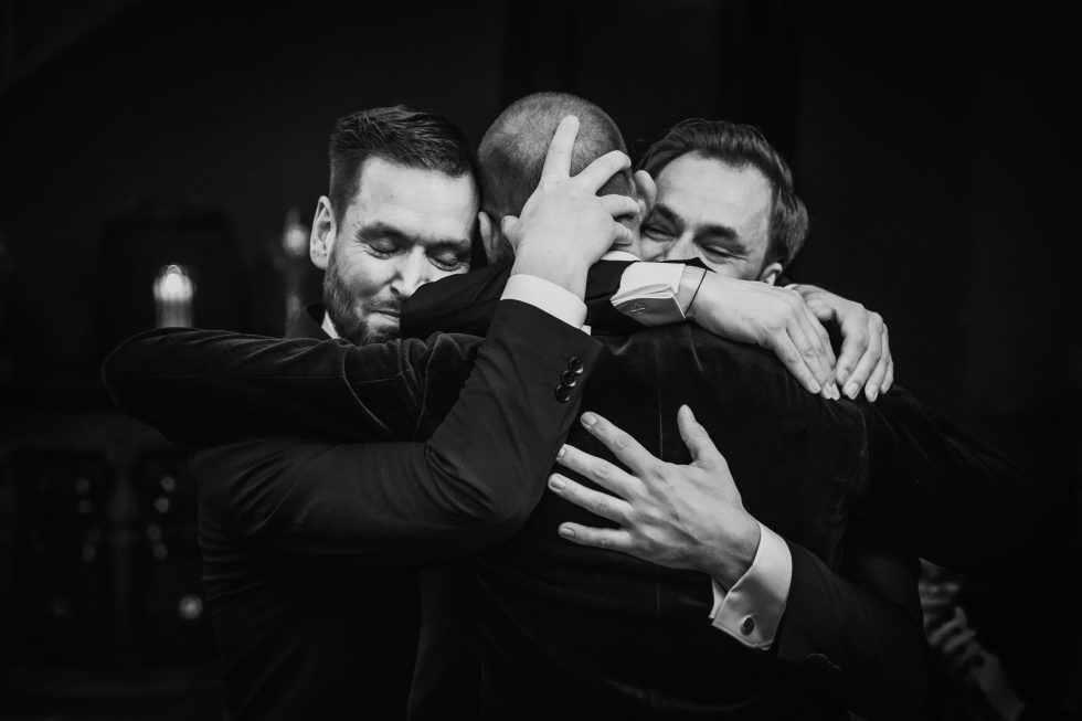 Emotionele omhelzing tussen drie mannen in smoking tijdens de bruiloft.