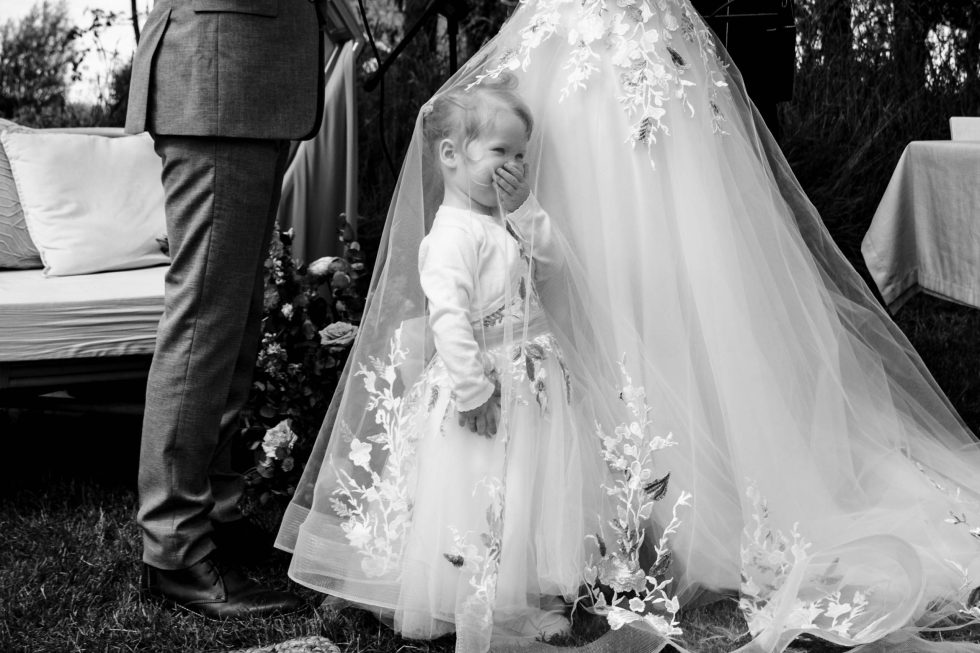 Klein meisje staat onder sluier van bruid en lacht verlegen tijdens ontroerende trouwceremonie.