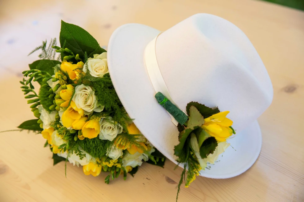 Bruidsboeket met gele en witte bloemen ligt naast een witte hoed met bloemcorsage op een houten tafel.