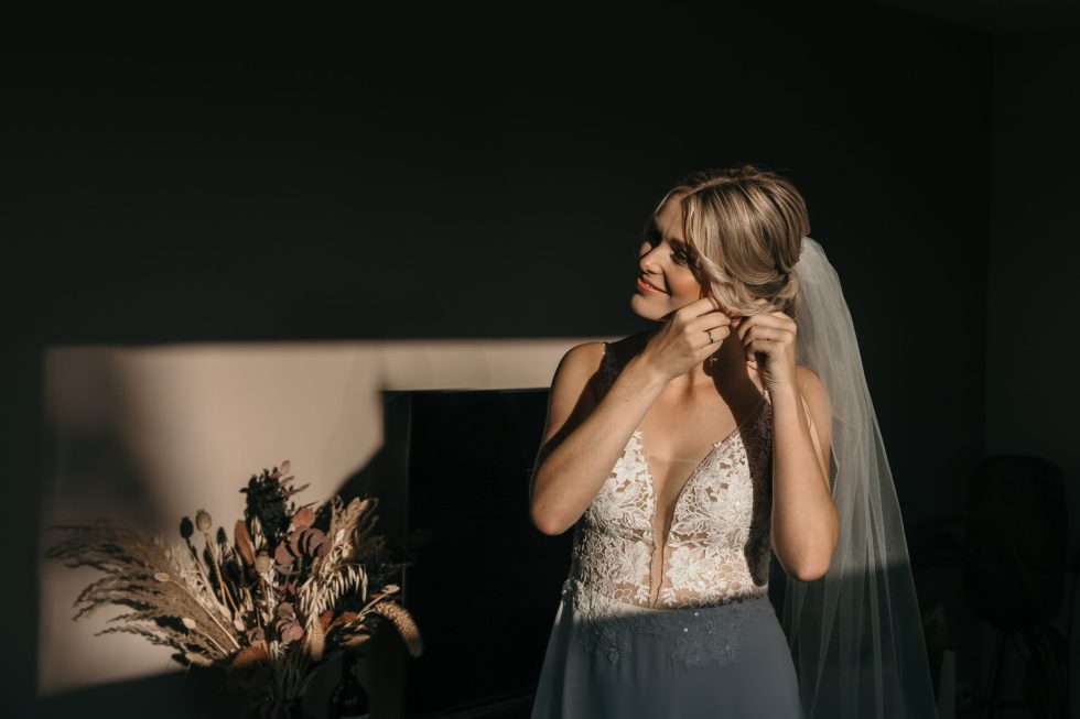 Bruid in kanten jurk doet oorbellen in terwijl zonlicht haar gezicht verlicht tijdens het klaarmaken op de trouwdag.