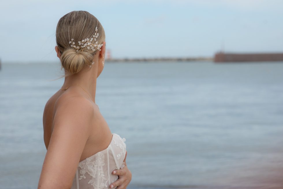 Bruid met laag opgestoken knot en een subtiele haarsieraad met parels en kristallen, staand aan het water in strapless kanten jurk.