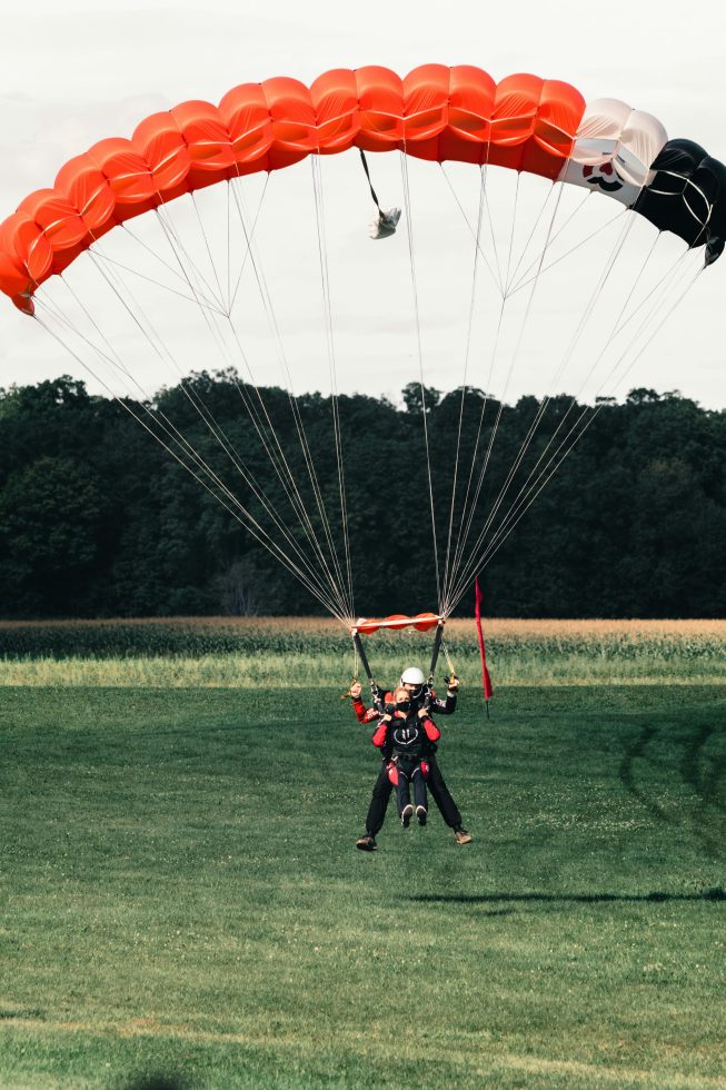 Twee mensen maken een tandemsprong met een rood-zwarte parachute en zijn net boven de grond aan het landen op een groot grasveld met bomen op de achtergrond.