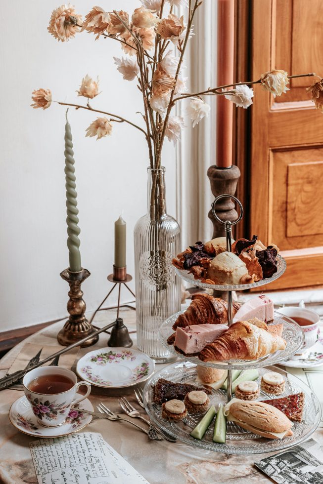 Gedekte tafel voor een high tea met een etagère vol croissants, sandwiches, gebakjes en snacks, omringd door vintage kopjes, kandelaars en een glazen vaas met gedroogde bloemen.