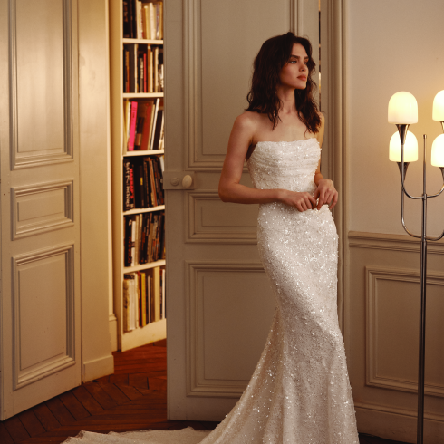 Bruid in een witte bruidsjurk die uitloopt vanaf de knie. De bruid poseert in een huis waar je een boekenkast ziet.