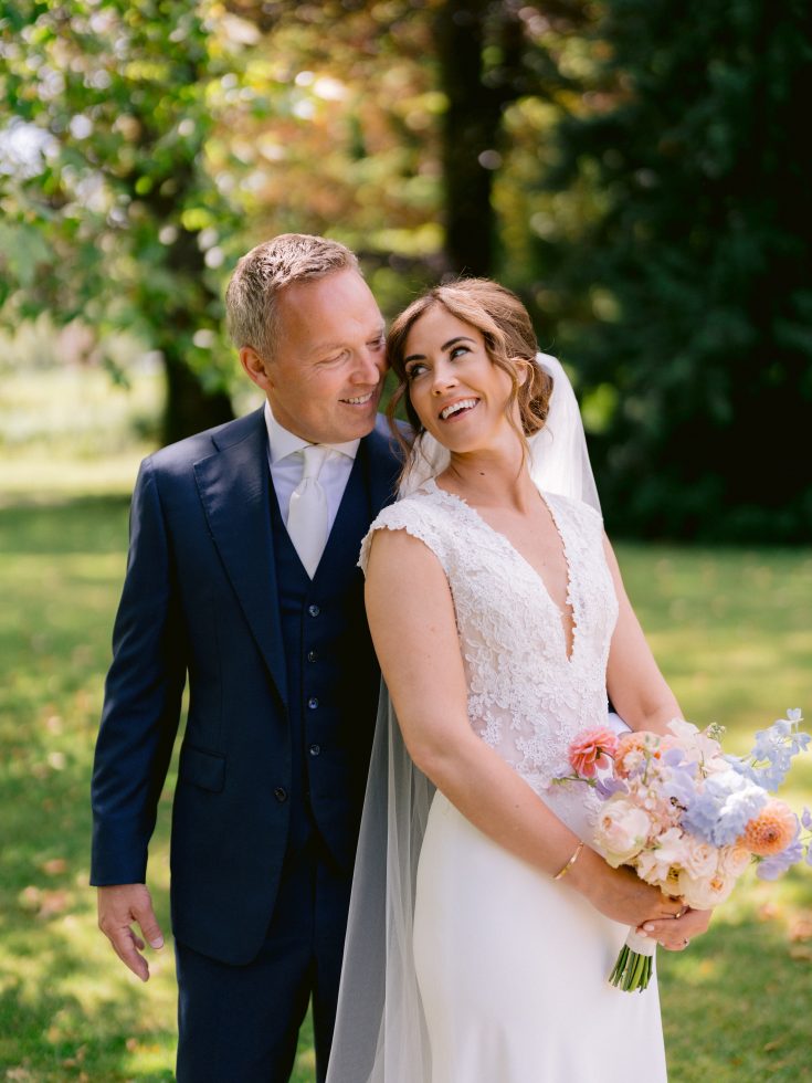 Een bruid en bruidegom poseren stralend samen in een zonnige, groene tuin. De bruid draagt een witte jurk met kanten details en houdt een kleurrijk boeket vast, terwijl de bruidegom in een donkerblauw pak liefdevol naar haar kijkt.