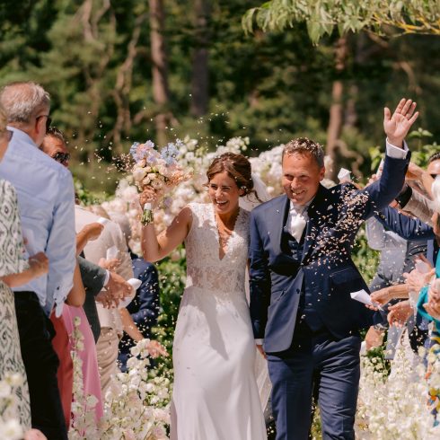 Een stralend bruidspaar loopt hand in hand over het bloemenpad terwijl gasten confetti gooien tijdens hun ceremonie op een zonnige dag in een groene buitenomgeving.