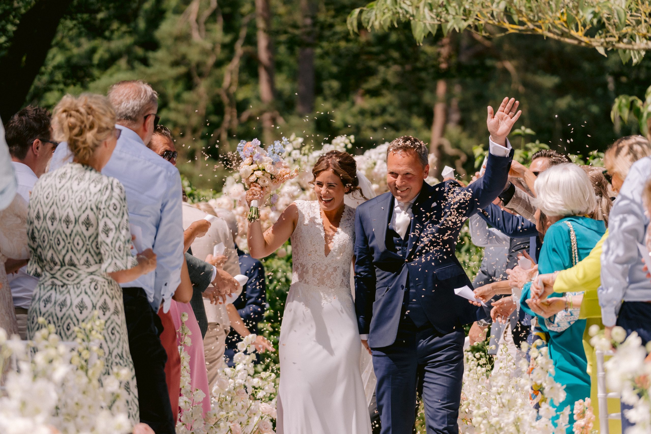 Een stralend bruidspaar loopt hand in hand over het bloemenpad terwijl gasten confetti gooien tijdens hun ceremonie op een zonnige dag in een groene buitenomgeving.