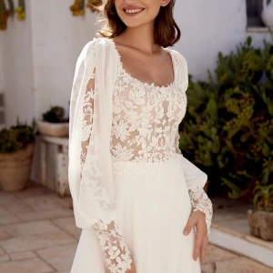 Vrouw in een elegante bohemian Lillian West trouwjurk met transparant kanten lijfje, pofmouwen en bloemenborduursels, staand op een zonovergoten terras met mediterrane beplanting.