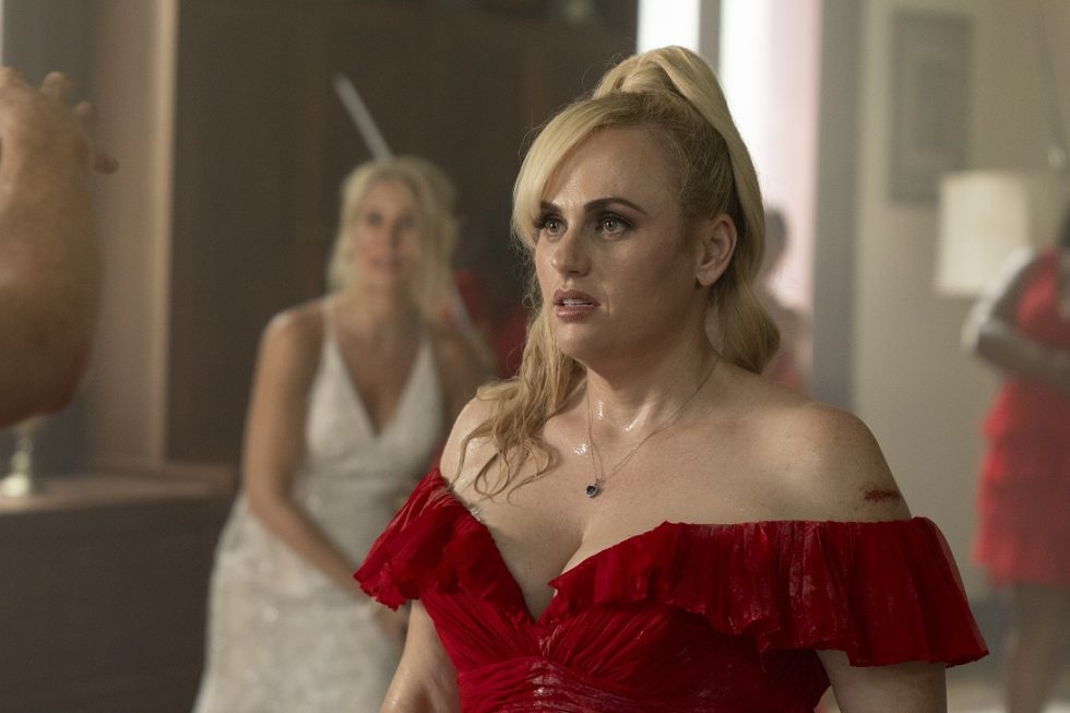 Rebel Wilson in een rode off-shoulder jurk, bezweet en met een bezorgde blik. Op de achtergrond staat een vrouw in een witte trouwjurk wazig in beeld, wat duidt op een gespannen scène.