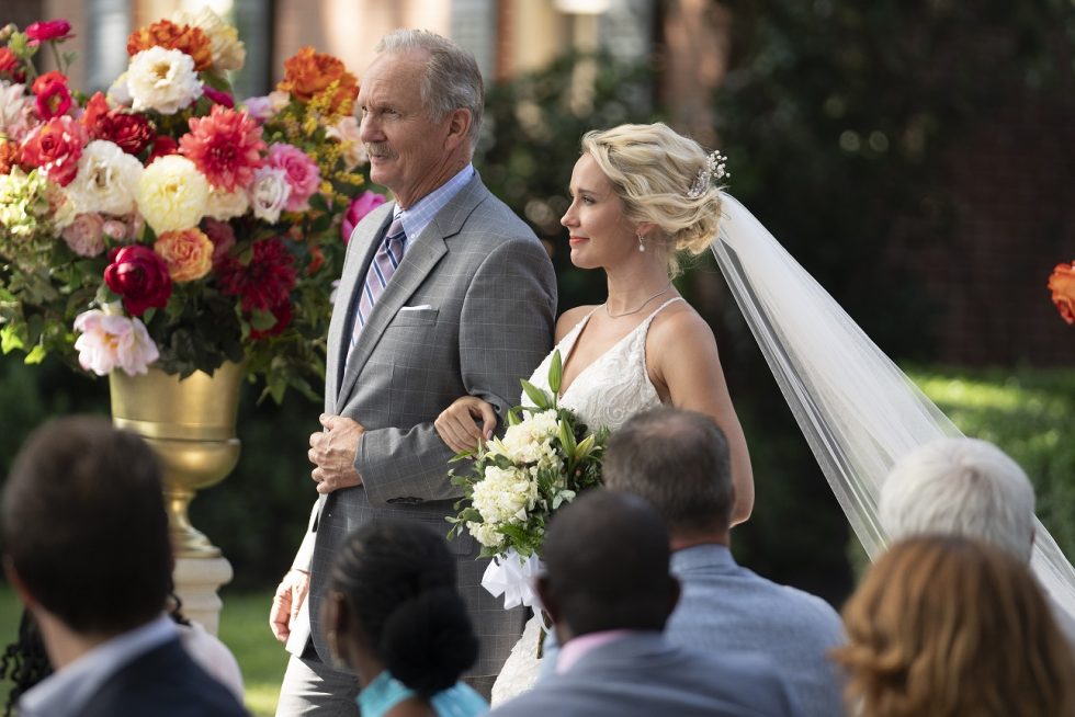 Een bruid in een witte jurk met sluier en bloemenboeket wordt naar het altaar begeleid door een oudere man in een grijs pak. Ze lopen langs zittende gasten, met kleurrijke bloemen op de achtergrond, tijdens een buitenceremonie.