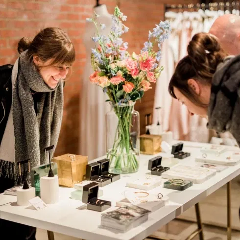 Bezoekers bekijken trouwringen en sieraden bij een stand op een bruidsbeurs, sfeervol gepresenteerd met bloemen en luxe displays.