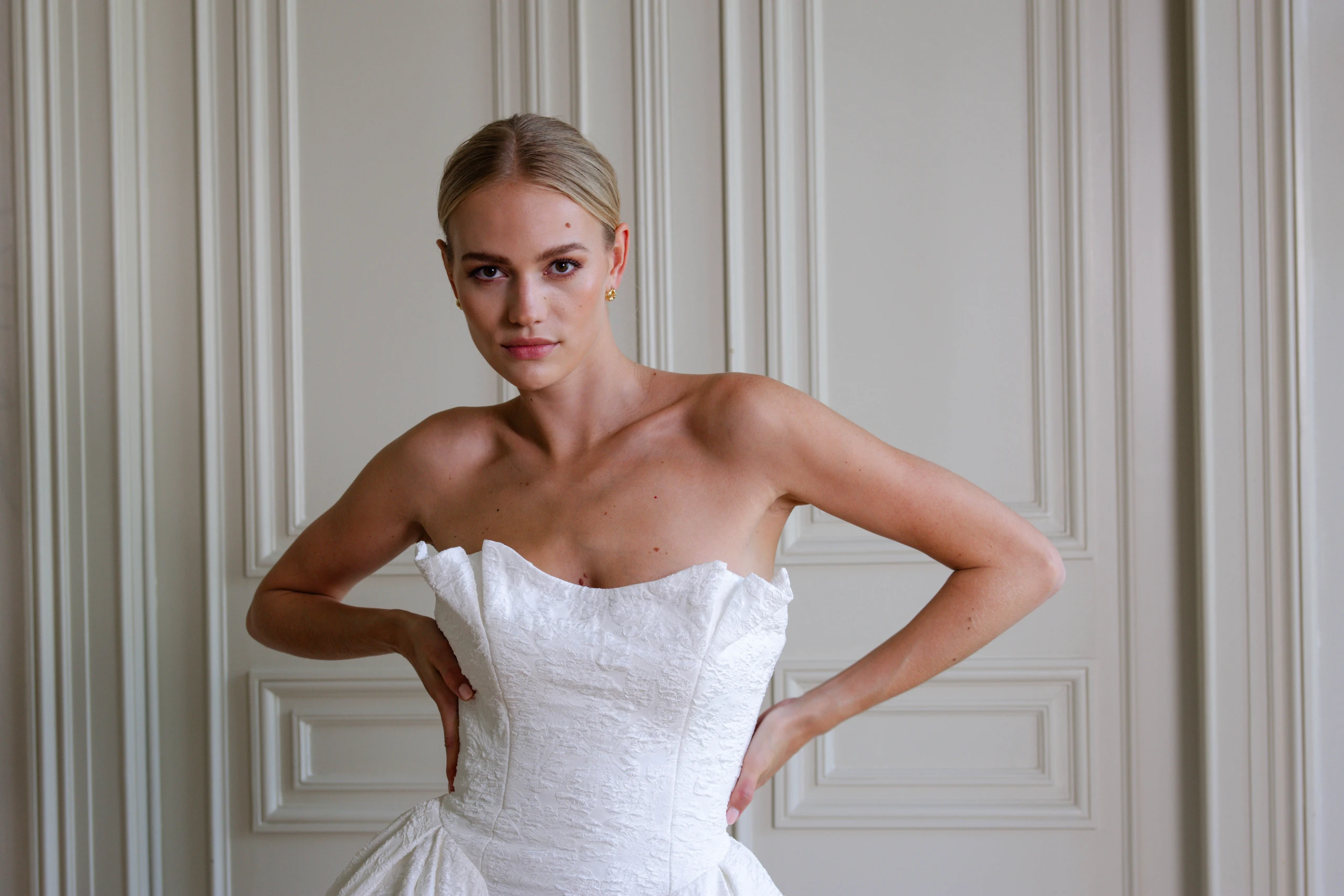 Bruid in een strapless witte trouwjurk met een elegante, gestructureerde top, poserend in een klassiek interieur met paneeldeuren.