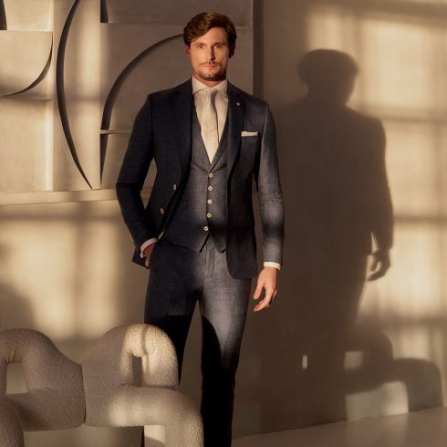 Man in stijlvol donkerblauw driedelig trouwpak met gilet en crèmekleurige stropdas, poserend in modern interieur met warme lichtinval van OFM.