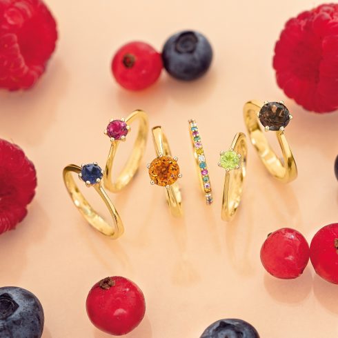 6 verschillende goude ringen met gekleurde diamanten op een rijtje. Eromheen liggen allemaal frambozen en blauwe bessen