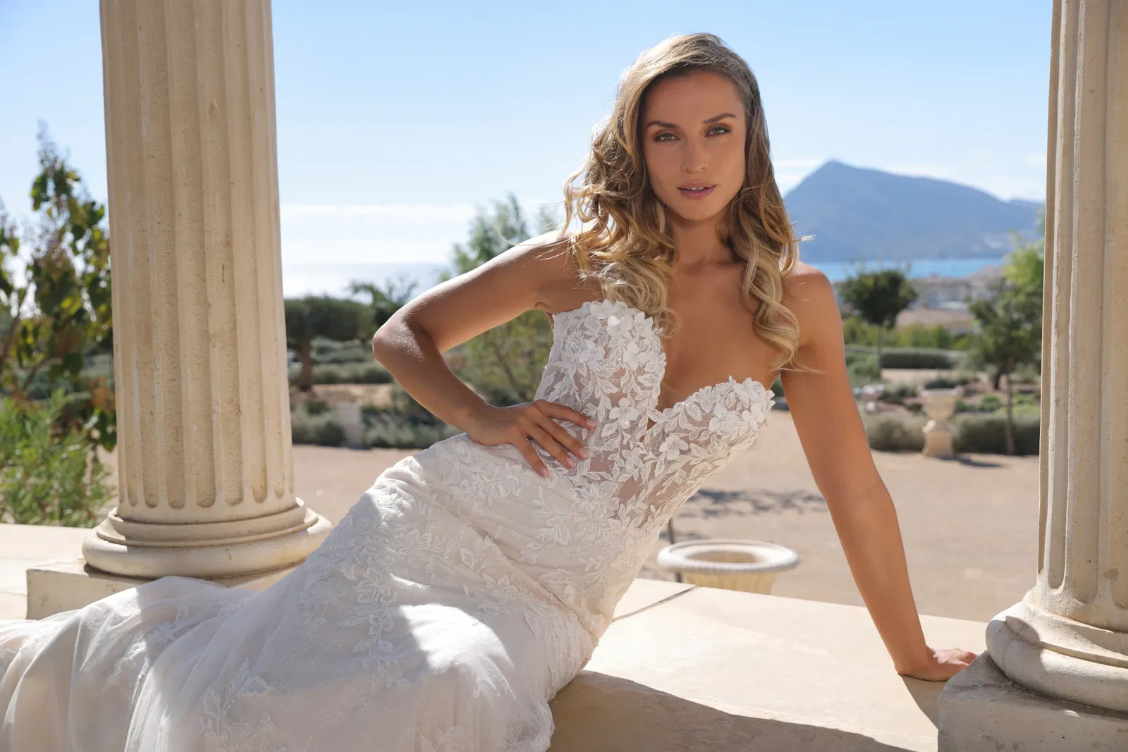Strapless bruidsjurk met sweetheart halslijn en kanten bloemendetails, gedragen door een bruid in een romantische buitenlocatie met klassieke zuilen en uitzicht op de zee en bergen.