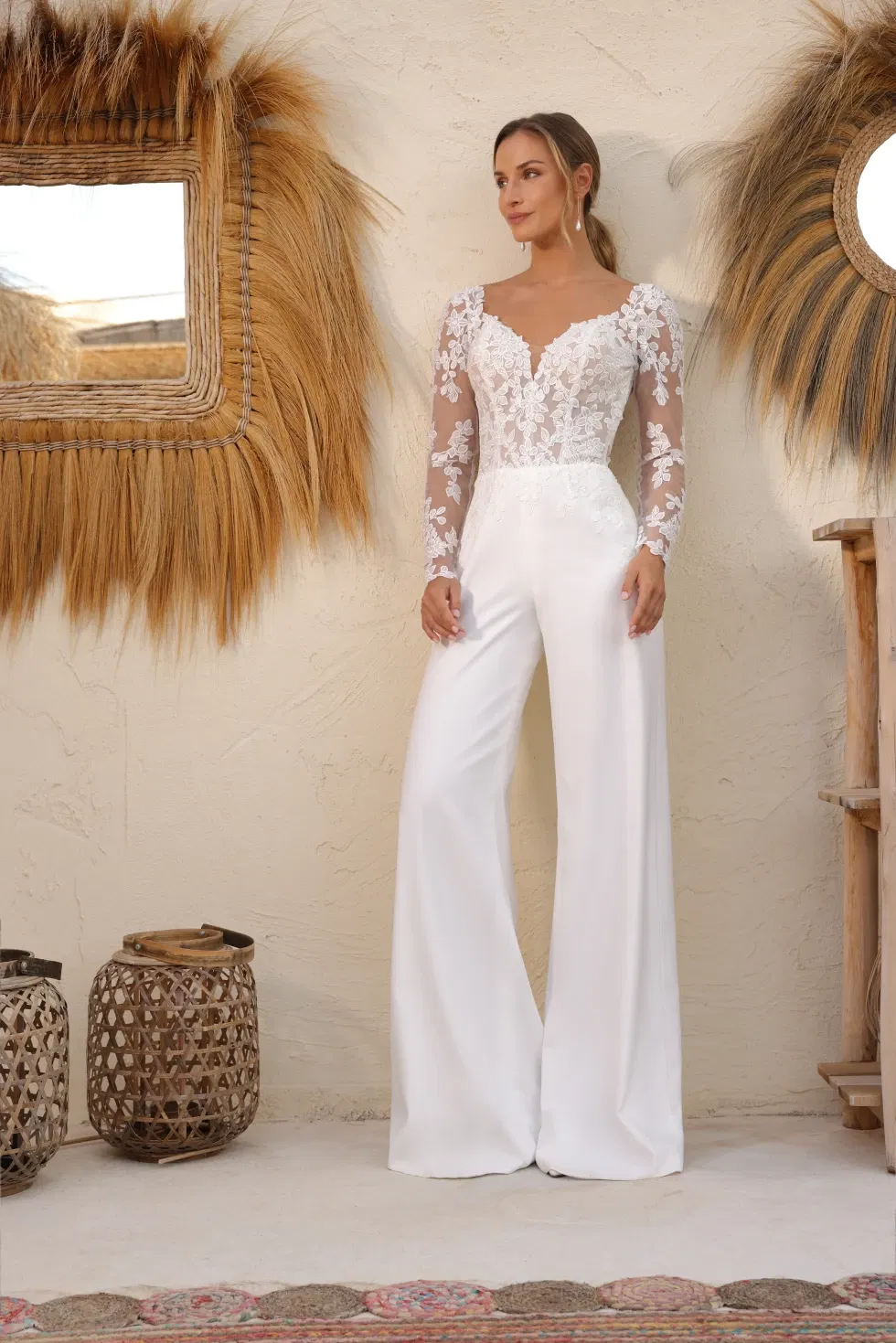 Een bruid draagt een moderne witte jumpsuit met wijde pijpen en een nauwsluitend lijfje van doorschijnende stof met kanten bloemenmotieven en lange mouwen. Ze staat in een stijlvolle, bohemian setting met natuurlijke decoratie.