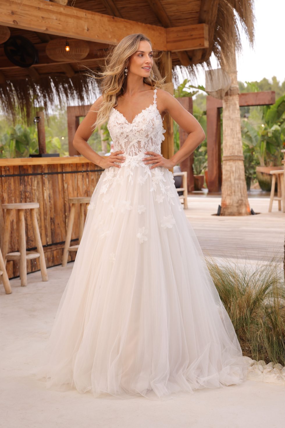 Romantische bruidsjurk met tule rok en kanten lijfje met bloemendetails en spaghettibandjes, gedragen door een bruid bij een tropische buitenlocatie met houten bar en palmen.