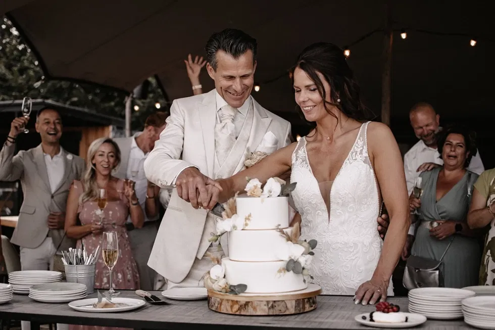 Bruid en bruidegom snijden samen een klassieke bruidstaart aan tijdens hun bruiloft, omringd door feestende gasten