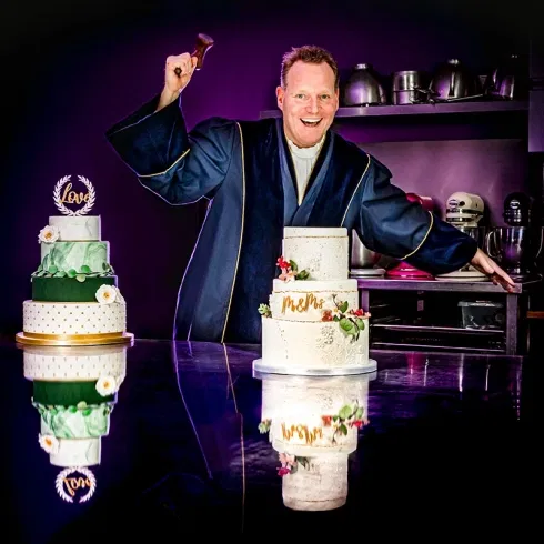 Trouwambtenaar in toga met twee bruidstaarten in kleurrijke bakkerijomgeving.