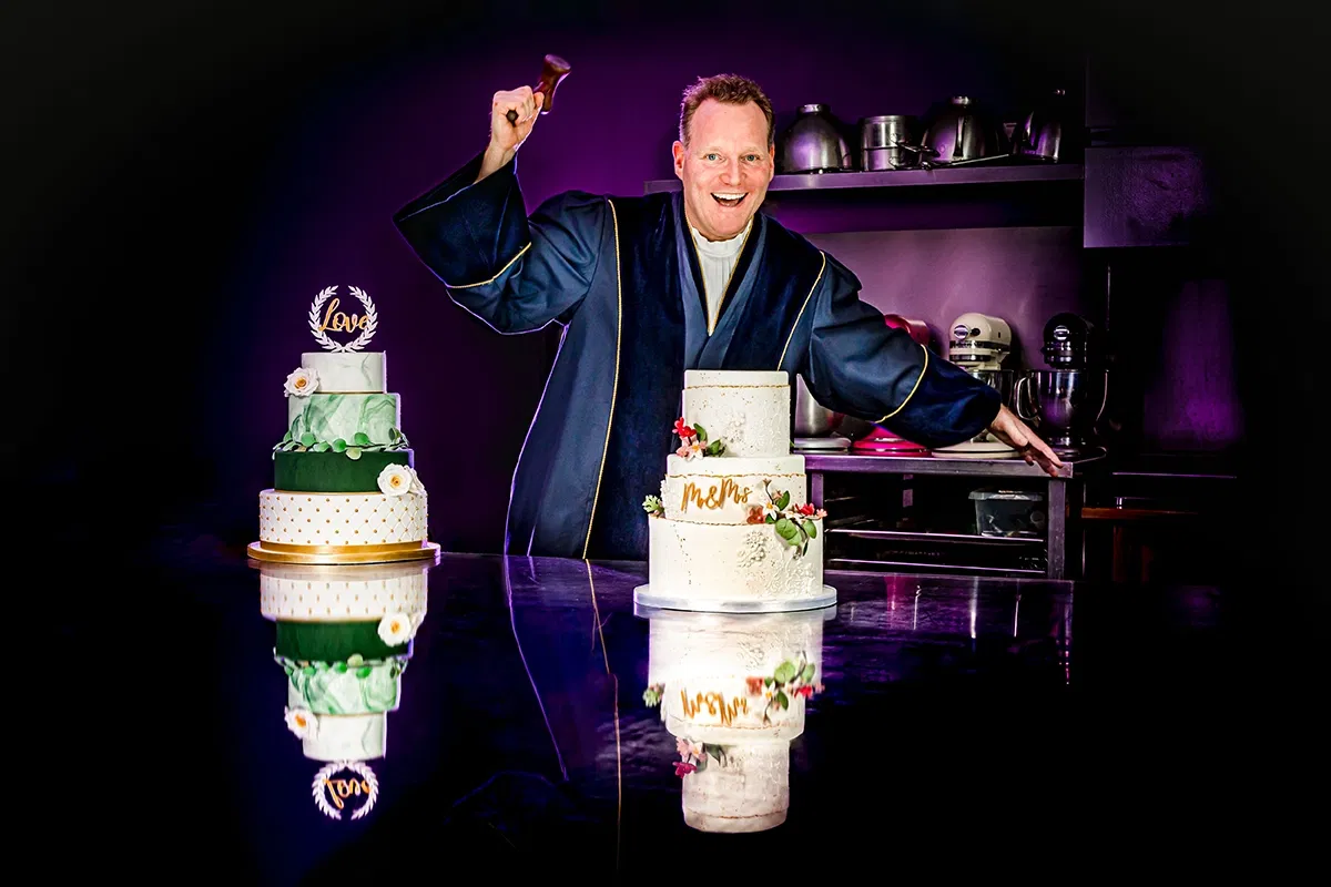 Trouwambtenaar in toga met twee bruidstaarten in kleurrijke bakkerijomgeving.