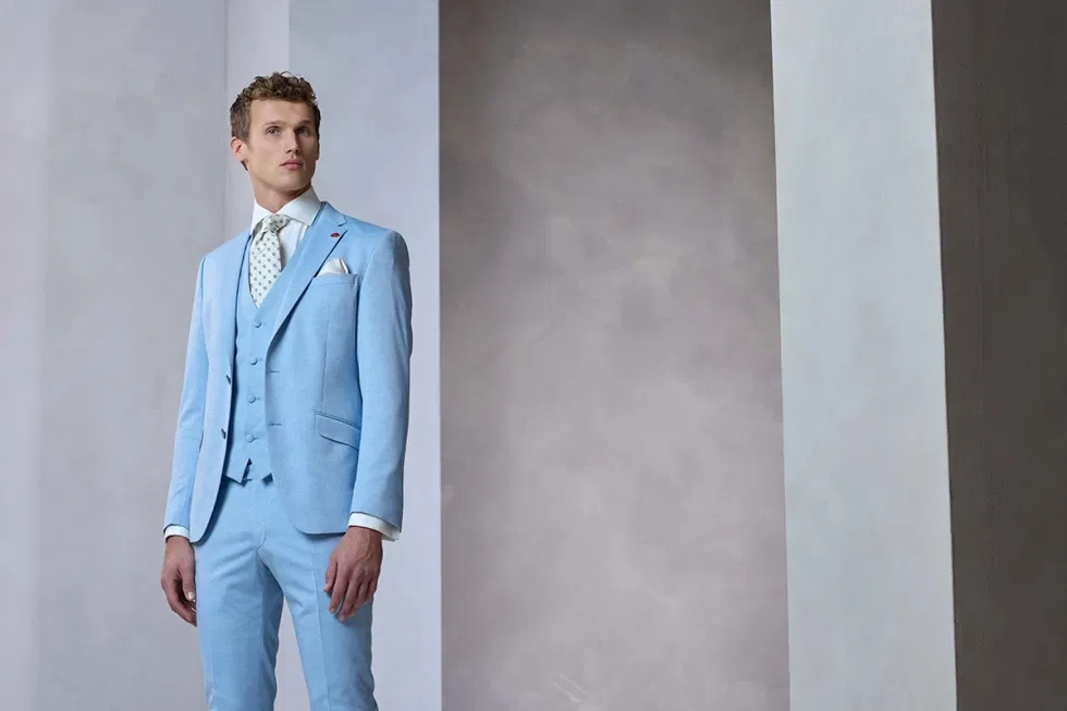 Man in lichtblauw driedelig trouwpak poseert stijlvol in een moderne, minimalistische ruimte.