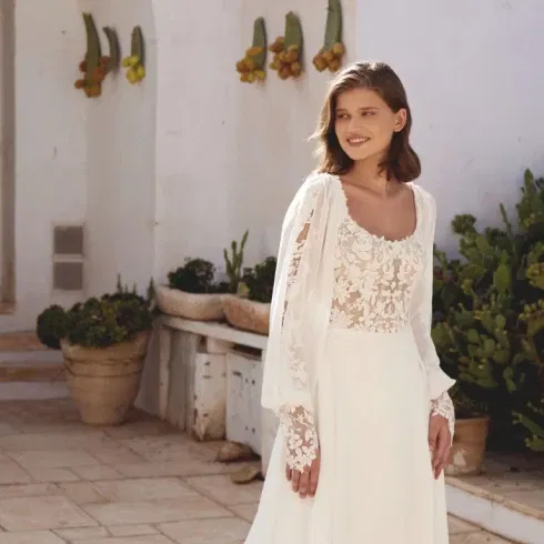 Vrouw in een elegante bohemian Lillian West trouwjurk met transparant kanten lijfje, pofmouwen en bloemenborduursels, staand op een zonovergoten terras met mediterrane beplanting.