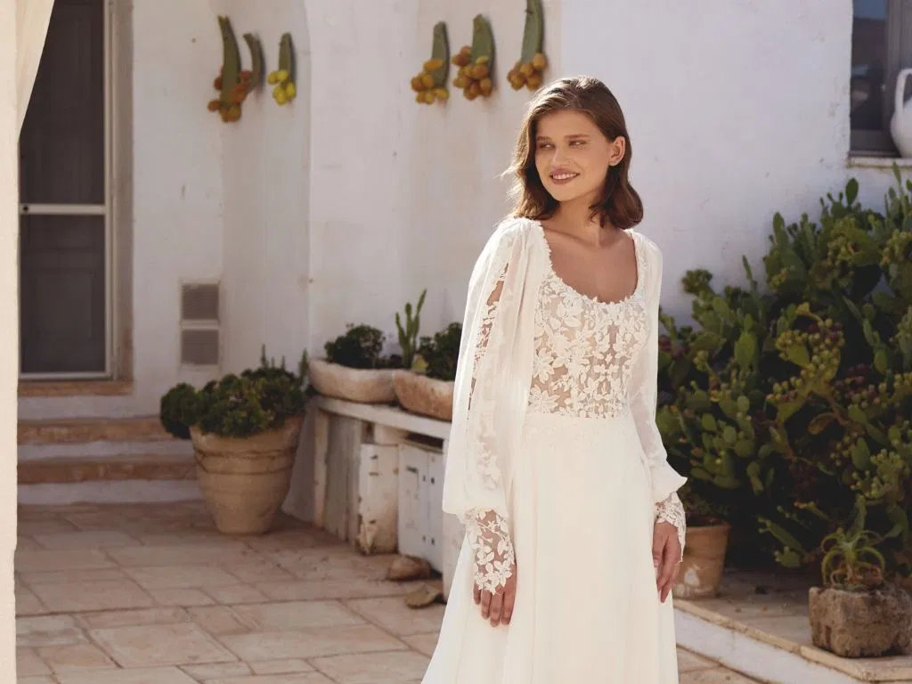 Vrouw in een elegante bohemian Lillian West trouwjurk met transparant kanten lijfje, pofmouwen en bloemenborduursels, staand op een zonovergoten terras met mediterrane beplanting.