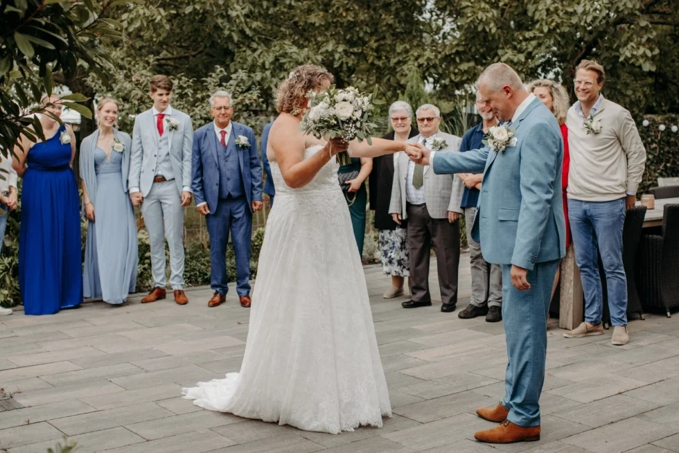 Bruid en bruidegom dansen hand in hand tijdens hun buitenbruiloft, omringd door lachende familie en vrienden.