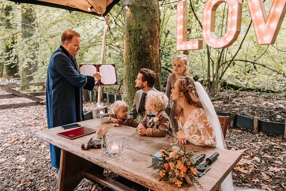Buitenbruiloft in het bos met trouwambtenaar en gezin aan tafel tijdens ceremonie.
