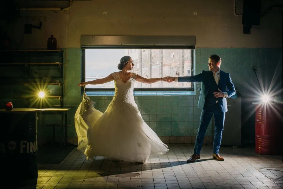 Bruid en bruidegom dansen sierlijk in een industriële setting met lichtspots op de achtergrond.