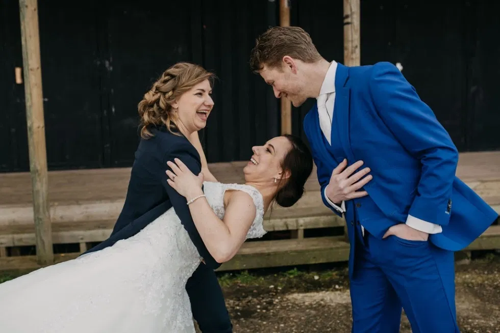 Twee vrouwen in trouwjurk en een man in blauw pak lachen samen tijdens een speelse trouwfoto.