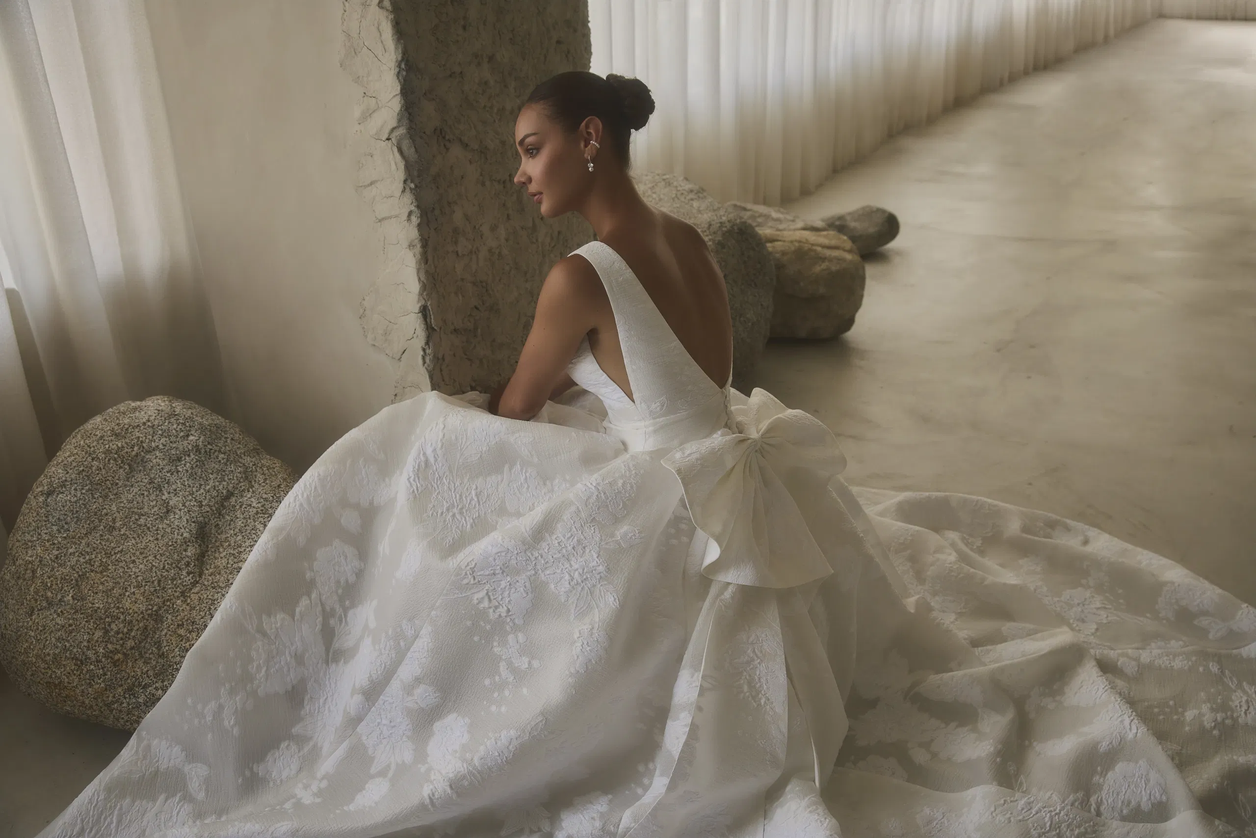 Romantische Casablanca trouwjurk met diepe V-rug, volumineuze rok en grote strikdetail, gefotografeerd in een elegante, minimalistische setting.