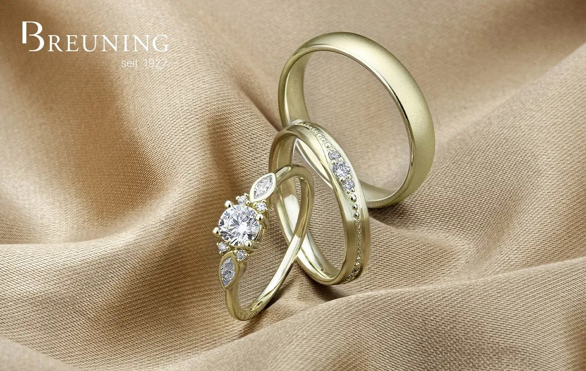 Drie gouden ringen, waaronder een verlovingsring met diamant en twee trouwringen, liggen op glanzende stof met een juwelierslogo in beeld