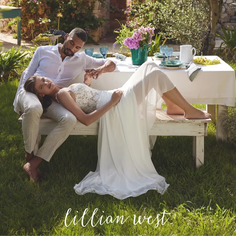 Een man en een vrouw zijn buiten aan een tafel. Hij zit rechtop en zij ligt op zijn schoot in een witte bruidsjurk van Lillian West