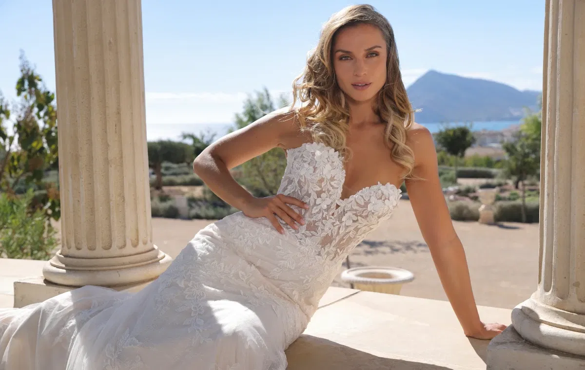 Strapless bruidsjurk met sweetheart halslijn en kanten bloemendetails, gedragen door een bruid in een romantische buitenlocatie met klassieke zuilen en uitzicht op de zee en bergen.